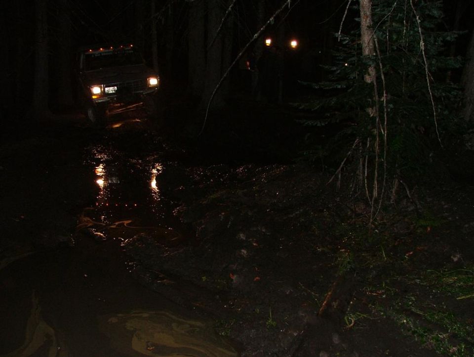 Front view of a dark-colored truck driving through a muddy area at night, with headlights on