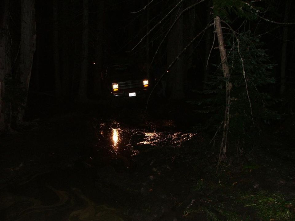 4x4 with headlights on facing a flooded mud hole on a dark forest trail at night