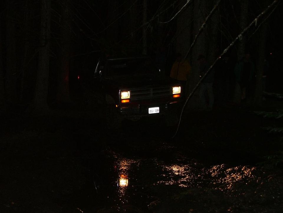 4x4 with headlights on at a night water crossing in the woods, onlookers watching