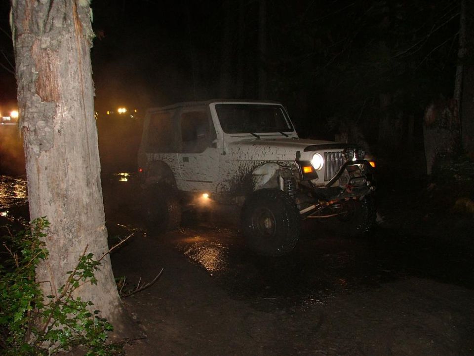 White Jeep Wrangler covered in mud, driving on a wet, dark trail at night. Another vehicle's lights are visible in the background