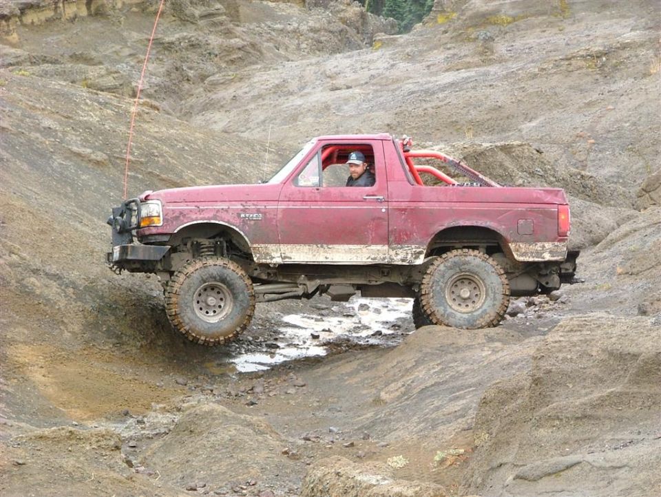 Red off-road truck with roll cage, lifted suspension, and large tires navigating a rocky, muddy terrain