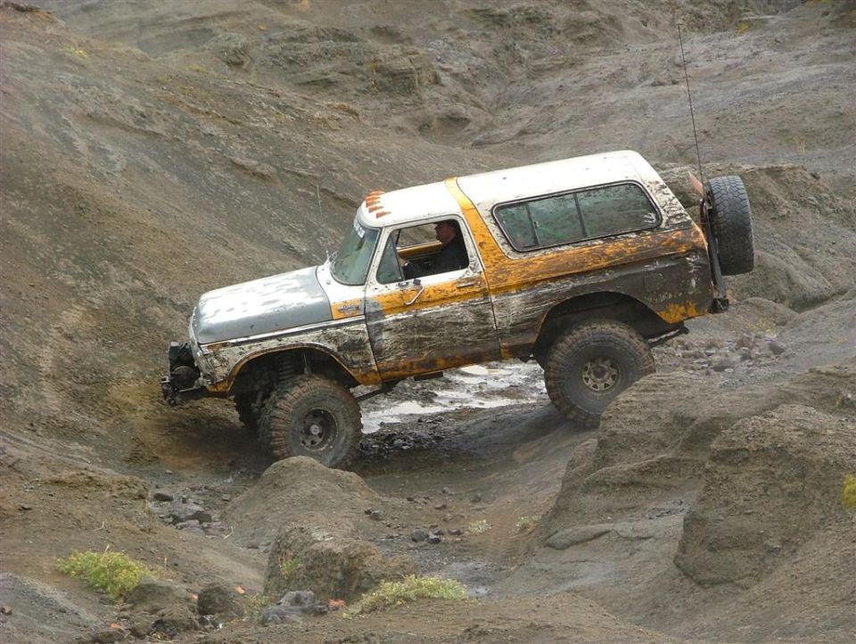 White and orange muddy Ford Bronco crawling over rocks in a gray wash, side view