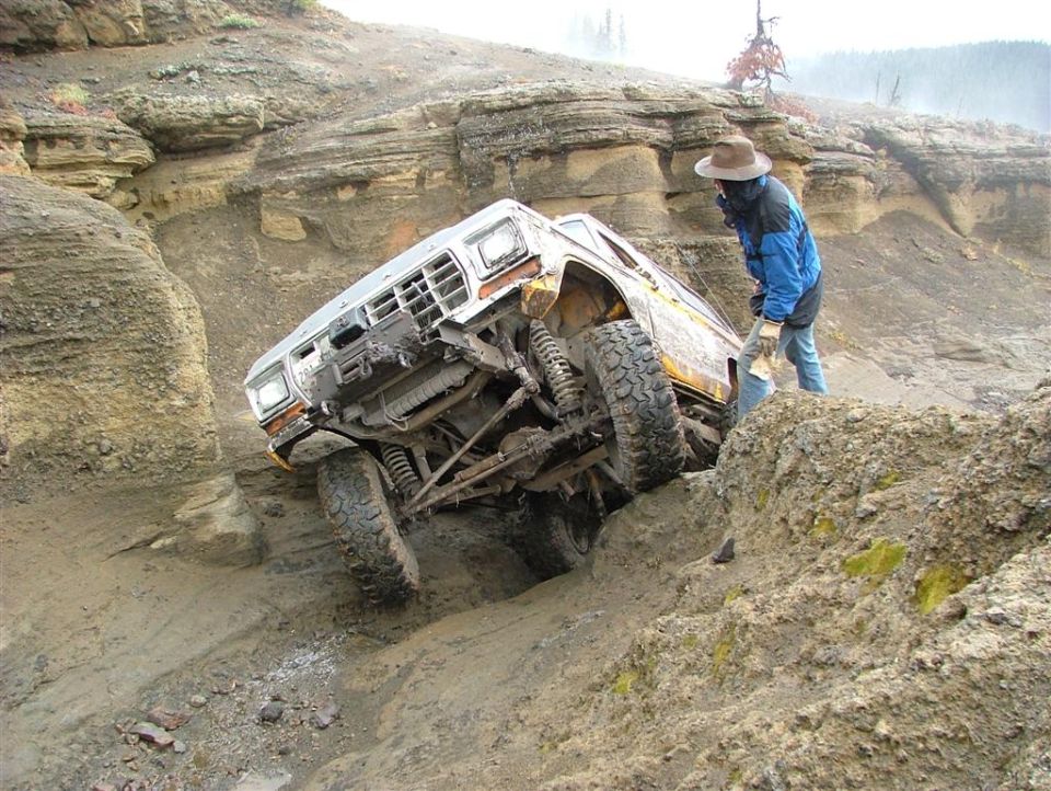 White off-road truck with lifted suspension climbing a muddy hill, driver in blue jacket and hat observing