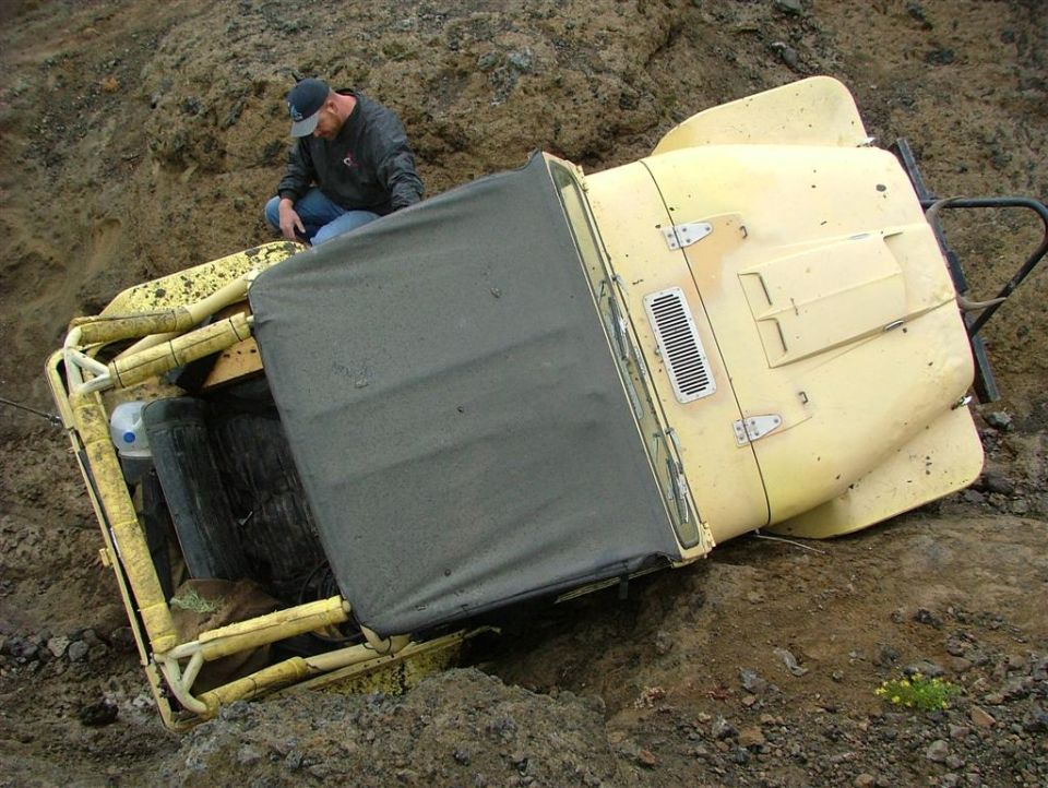 Yellow off-road vehicle tipped on its side in dirt, with a man looking at it