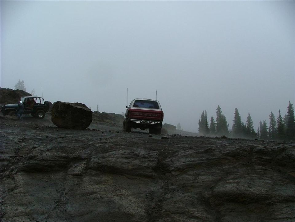 Two off-road vehicles driving on a rocky, foggy mountain trail. One is a red truck, the other is a dark-colored Jeep