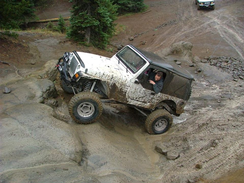 White Jeep Wrangler navigating a muddy, rocky trail, driver visible