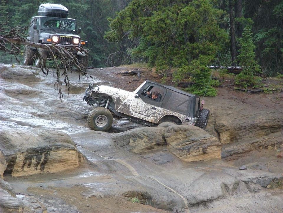 White Jeep Wrangler navigating a rocky, wet trail; another Jeep follows