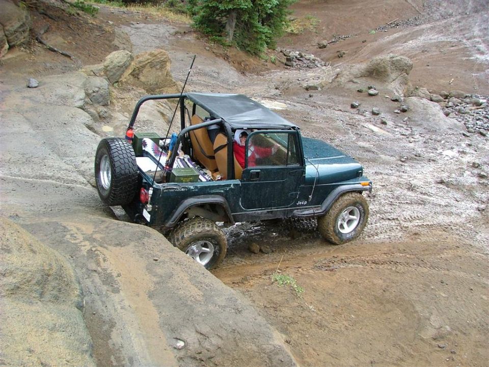 Green Jeep Wrangler crawling over rocks and mud, rear 3/4 view