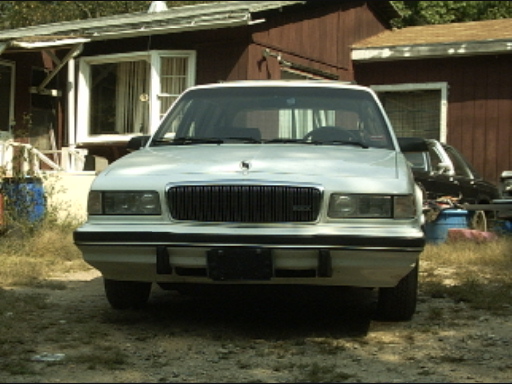 Front view of a white sedan with a black grille, parked outdoors. Another dark vehicle is visible behind it