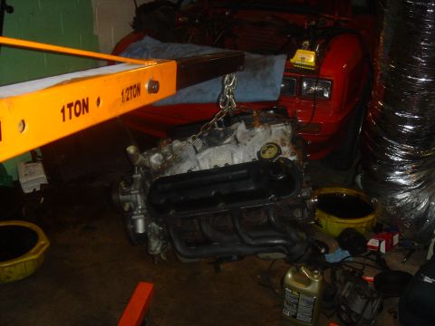 1986 Ford Mustang - An engine hoist is lifting a V8 engine, with a red car visible in the background.