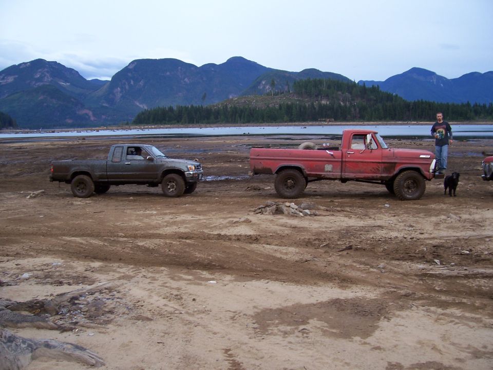 Side view of a gray pickup truck and a red pickup truck in a muddy area with mountains in the background