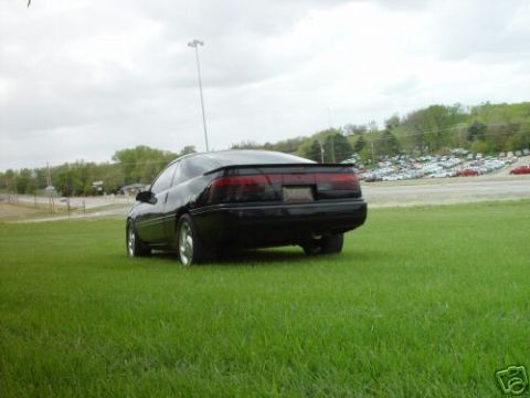 1992 Ford Probe - A black coupe is parked on a grassy field with a parking lot full of cars in the background.