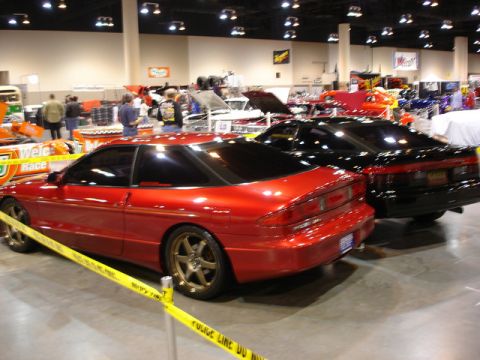 1992 Ford Probe - A red sports coupe is parked next to a black car at an indoor event, with yellow caution tape in the foreground.