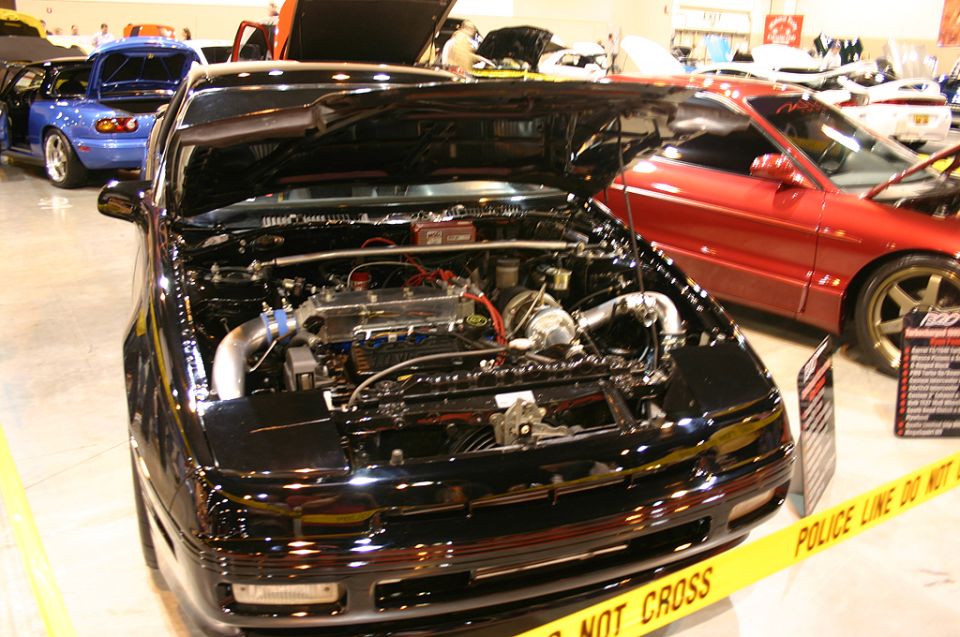 Black sports car, front view, hood open revealing engine, yellow police line tape in foreground