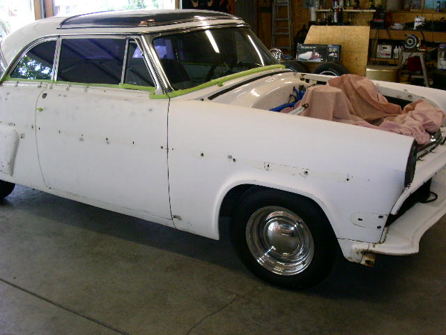 White 1950s Ford coupe mid-restoration in a garage, hood off with engine bay covered.
