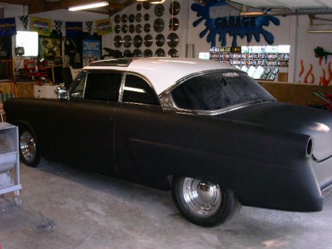 1954 Ford Crestline - A customized coupe with a matte black finish and a white roof is shown in a garage setting with various license plates displayed on the wal…