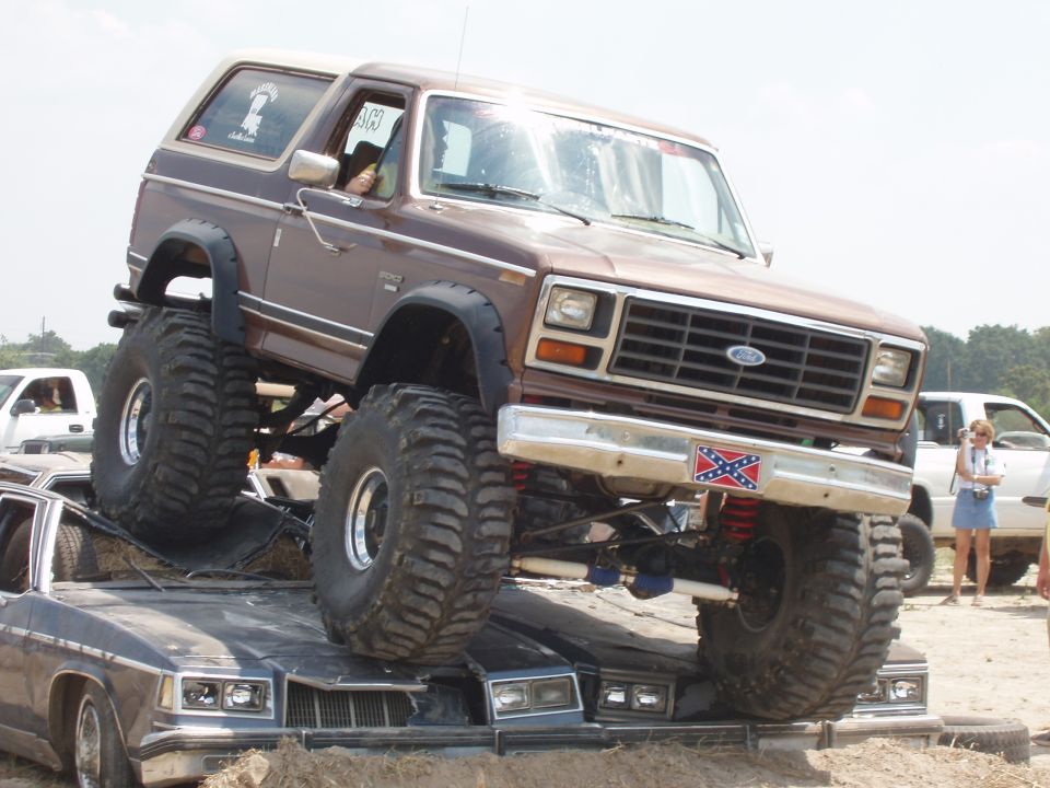 Brown lifted Ford Bronco on top of two crushed cars, with a Confederate flag decal on the bumper