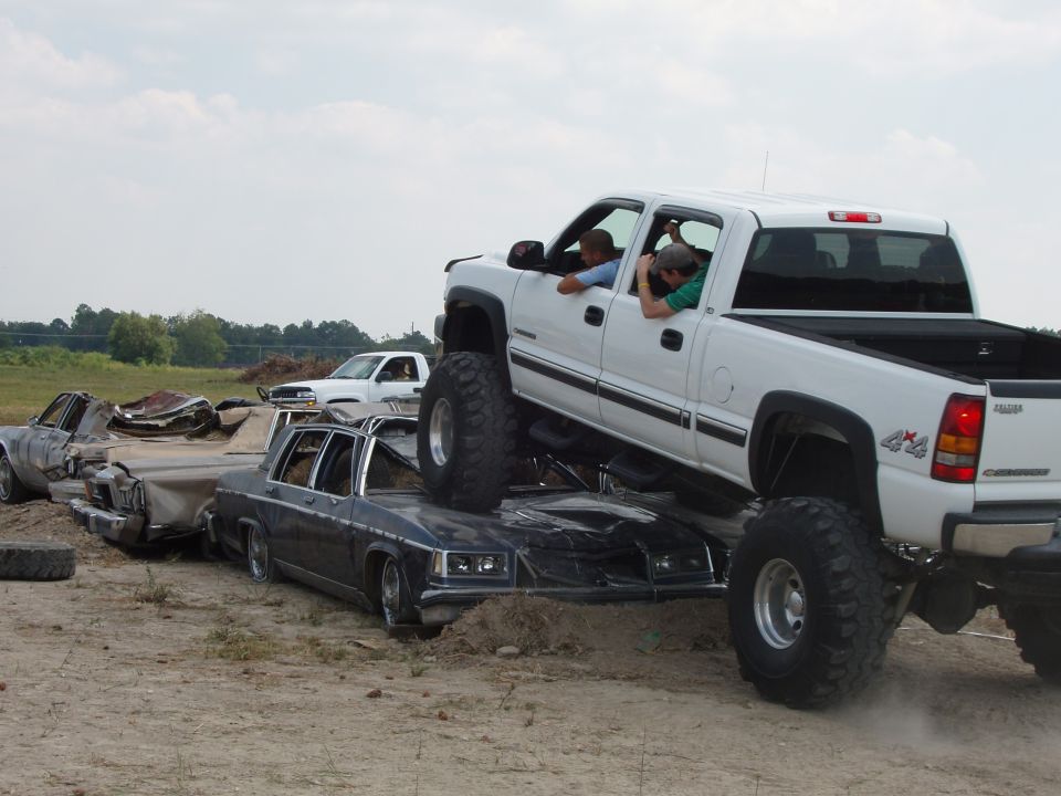 Lifted white pickup truck on top of a dark sedan, with another white pickup and a wrecked car in the background