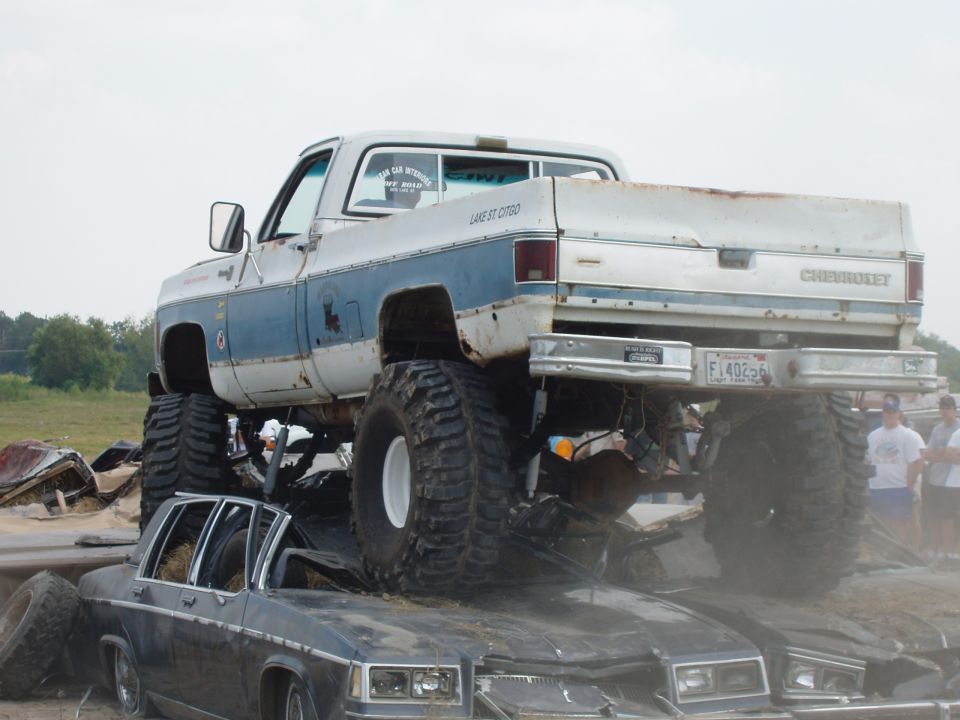 White and blue lifted Chevrolet pickup truck with large tires crushing a dark sedan, rear view