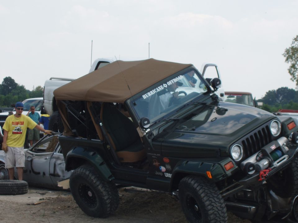 Green Jeep Wrangler TJ with winch and mud tires crawling over a crushed car