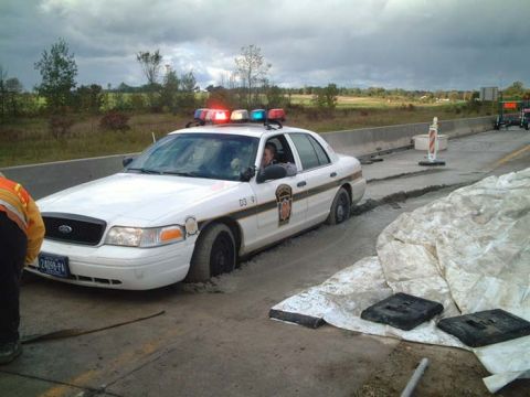1972 Ford Ranchero - A police car is stuck in wet concrete on a highway construction site, with a worker nearby.
