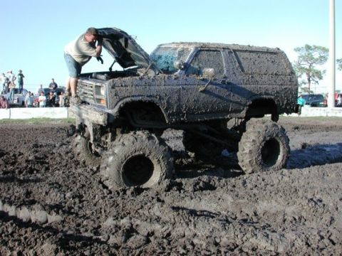 1979 Ford Bronco - A man checks the engine of a heavily mud-covered SUV stuck in a mud pit, with spectators in the background.
