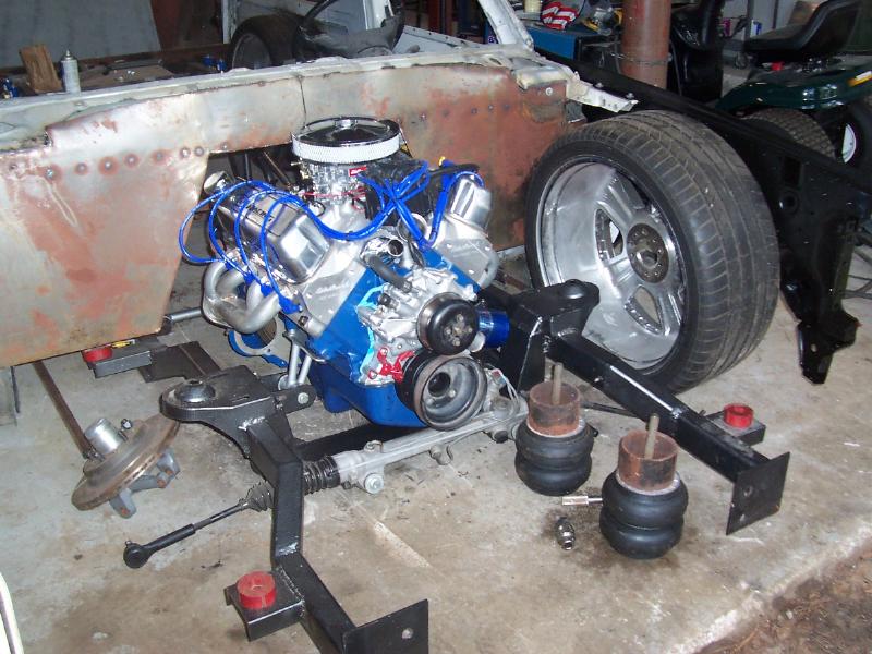 Engine assembly with blue wiring, air intake, and exhaust headers, next to a large tire and suspension components