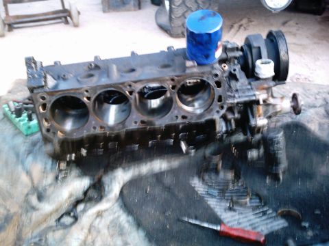 1986 Ford Bronco - A bare V8 short block sits on a garage floor with the cylinder heads removed, exposing four honed bores on the near bank and the deck surfa…