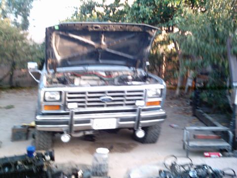 1986 Ford Bronco - A head-on view of a light gray/primer 1980s Ford truck with the hood raised, revealing an engine bay mid-work with red plug wires visible.…