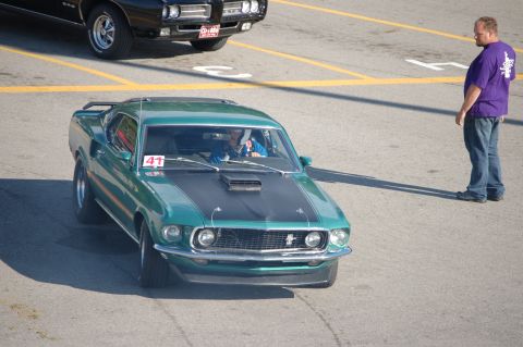 2005 Ford Deluxe - A man in a purple shirt observes a green 1969 Ford Mustang Mach 1 as it sits on asphalt, with another car in the background.
