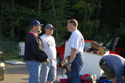2005 Ford Deluxe - People gathered around a trailer with a red car in the background, possibly at an event.
