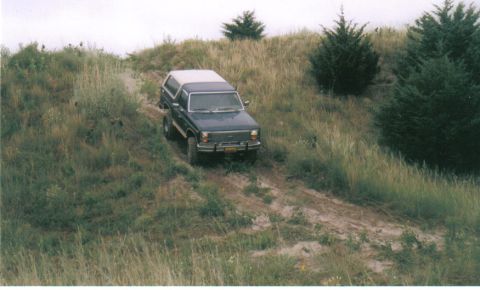 1986 Ford Bronco - A dark blue 1986 Ford Bronco with a white top navigates a dirt path on a grassy incline, surrounded by trees.