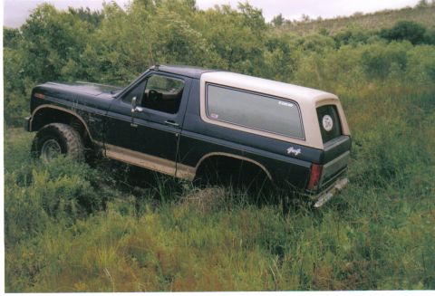 1986 Ford Bronco - Elevated three-quarter rear view of a dark blue and tan full-size Ford Bronco sitting nose-down and bogged in a muddy rut in tall grass and…