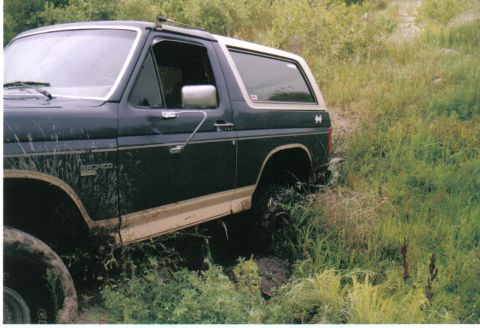 1986 Ford Bronco - Scanned side view of a dark green and tan Ford Bronco sitting down in a muddy rut surrounded by tall weeds, with mud spray up the doors and…