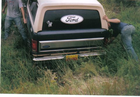1986 Ford Bronco - Scanned rear view of a dark green and tan two-tone Ford Bronco bogged down in tall grass, with two people pushing on the tailgate and rear…