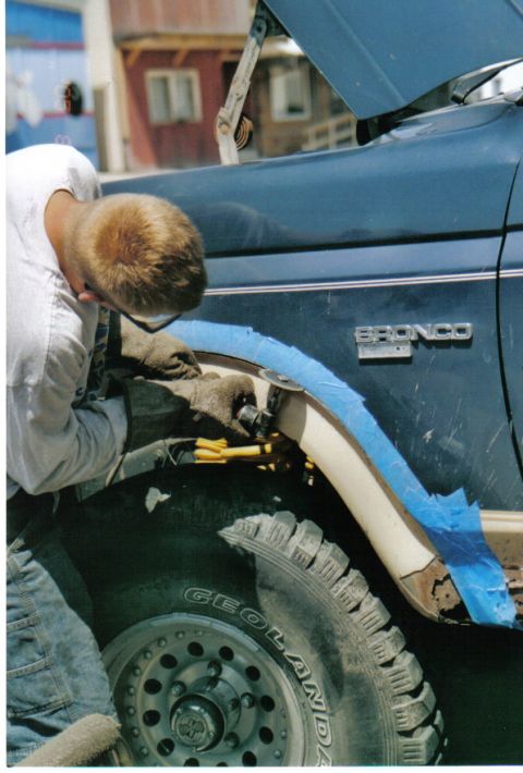 1986 Ford Bronco - A person modifies the fender flare of a blue 1986 Ford Bronco, with the 'BRONCO' badge clearly visible on the side.