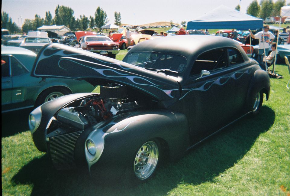 Satin black flamed 1930s coupe street rod with hood up at a grassy car show