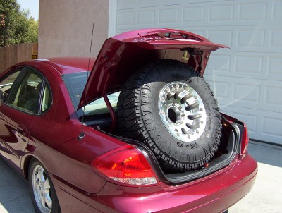 1995 Ford Bronco - A large off-road spare tire is stored in the trunk of a red sedan.