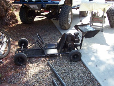 1995 Ford Bronco - A black go-kart sits on gravel, with a large truck partially visible in the background and a bicycle wheel to the left.