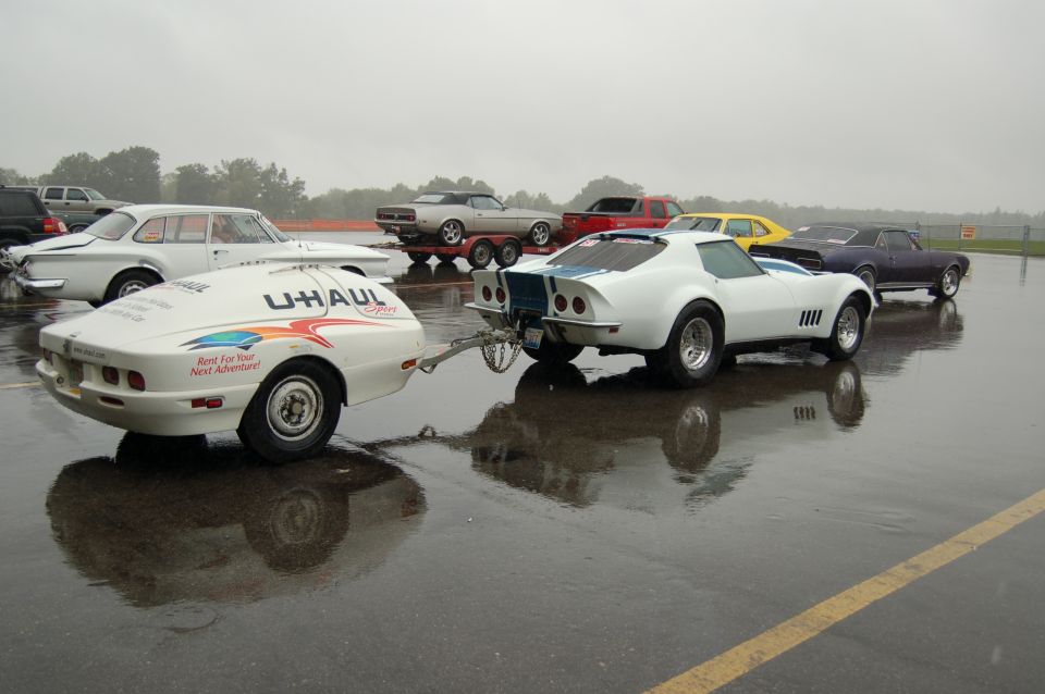 White C3 Corvette towing a small U-Haul Sport trailer in a rain-soaked parking lot with other cars