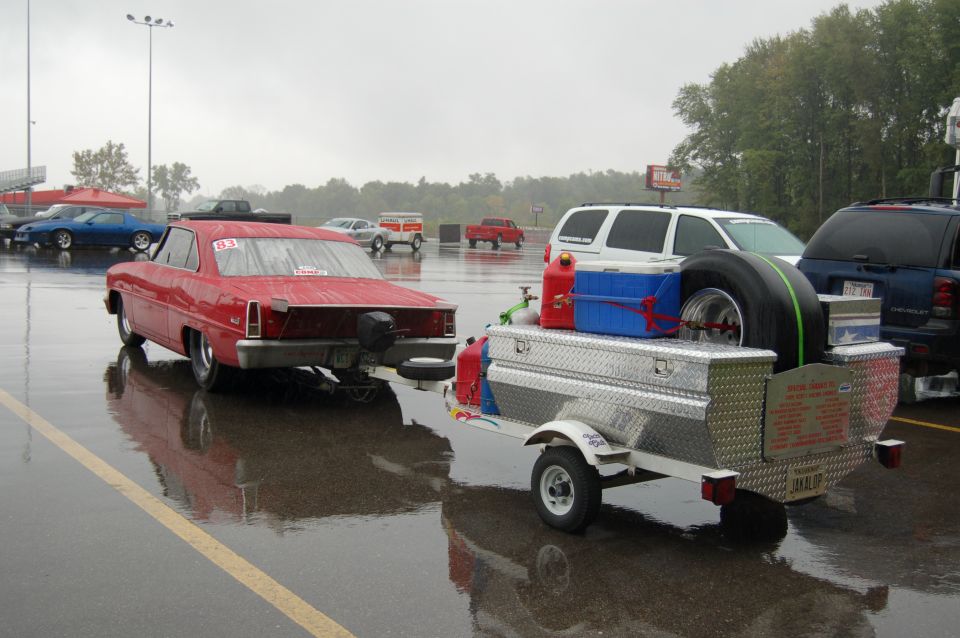 Red drag race coupe #83 towing a diamond-plate trailer loaded with a slick tire and cooler on a wet track pit area