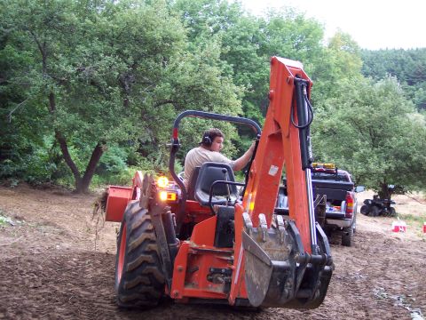 2001 Ford Crown Victoria - A person operates an orange tractor with a backhoe attachment in a wooded area, with a pickup truck and ATV visible in the background.