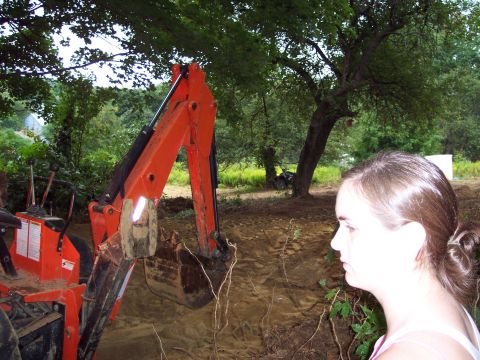 2001 Ford Crown Victoria - A woman observes an orange backhoe loader in a dirt area, with trees and a field in the background.