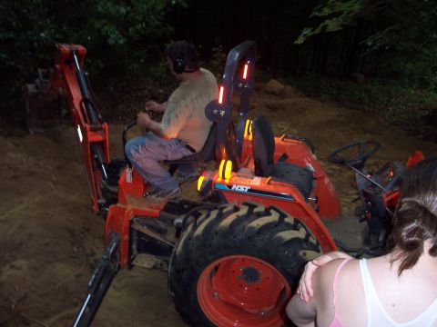 2001 Ford Crown Victoria - A man operates a tractor with a backhoe attachment in a wooded area, with another tractor and person nearby.