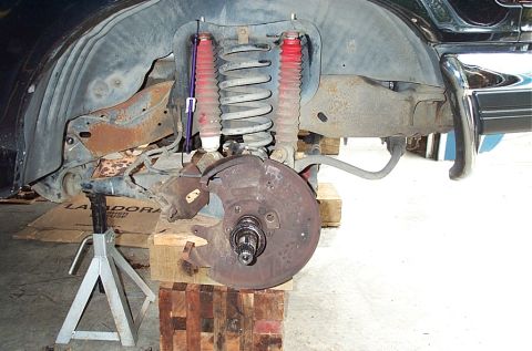 1995 Ford Bronco - The front corner of a dark-blue vehicle raised on a jack stand and wood blocks with the wheel removed, exposing the coil spring, twin shock…