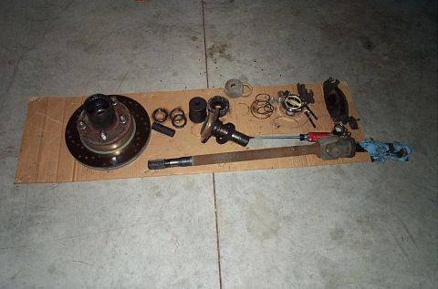 1995 Ford Bronco - A disassembled front axle laid out on cardboard on a garage floor: rotor and hub assembly with wheel studs at left, spindle bearings, snap…