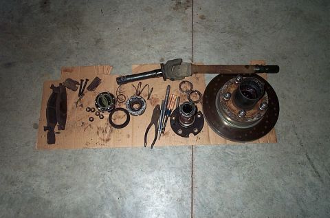 1995 Ford Bronco - Overhead shot of a front axle teardown spread on cardboard: the rotor-and-hub with wheel studs at right, a splined axle shaft with U-joint…