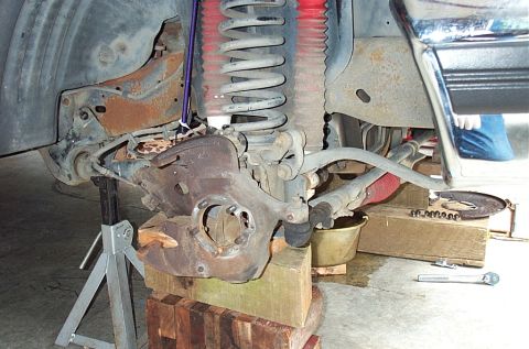 1995 Ford Bronco - Wider view of a front-end rebuild on a lifted truck: the bare steering knuckle sits on wood cribbing with the spindle and hub removed, next…
