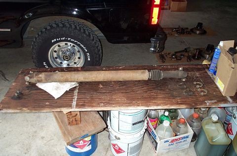 1995 Ford Bronco - A removed axle tube/shaft assembly with a rubber boot rests on a plank workbench in a garage, with U-joint caps and a shop rag beside it an…