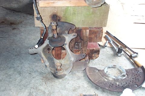 1995 Ford Bronco - A removed front steering knuckle stands on the garage floor against wood cribbing, with its upper and lower ball joints and threaded studs…